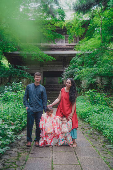 A family of four stands on a stone path surrounded by lush greenery, with two children in traditional kimonos, and a temple structure in the background.