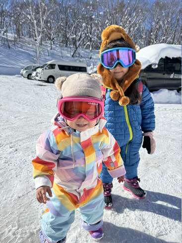 Two young children dressed in colorful snowsuits and goggles enjoy a snowy day. Behind them, cars and snow-covered trees create a bright, wintery background.