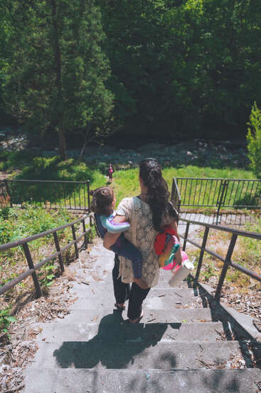 A woman carrying a small child descends outdoor stone steps, surrounded by lush greenery and trees, heading towards a river or stream below. Bright daylight shines.