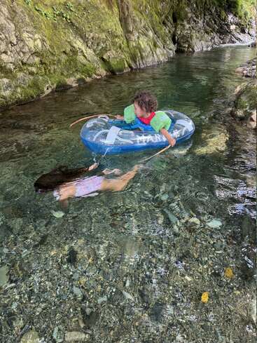 Two children enjoy a clear, rocky river; one floats in an inflatable ring, the other swims underwater. The scene is natural, peaceful, and adventurous.