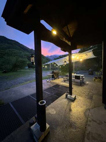 Evening scene with glowing string lights, a covered patio, picnic tables, mountains in the distance, lush greenery, and a purple-pink sunset sky, creating a peaceful atmosphere.