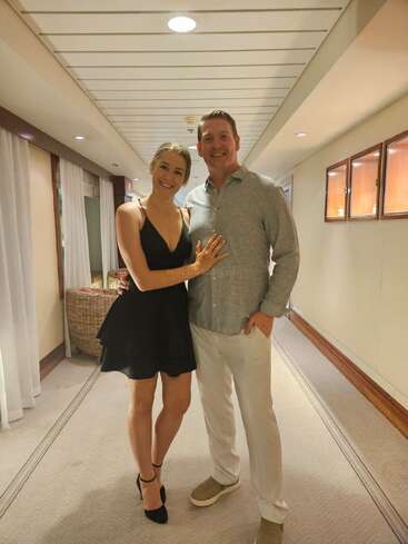 A smiling couple poses together in a hallway. The woman wears a black dress and heels; the man wears a grey shirt with light-colored pants and shoes.