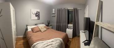 A cozy bedroom with a double bed, brown bedding, pillows, wall art, wardrobe, TV, nightstand, lamp, air conditioner, curtains, and wooden flooring.