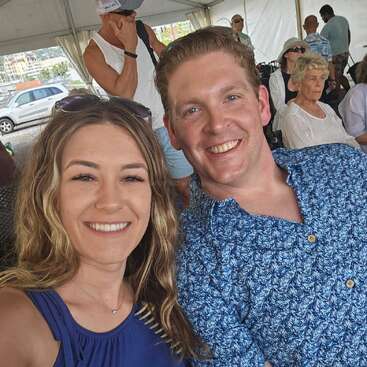 A smiling man and woman take a selfie under a tent. Several people sit and stand in the background. Everyone appears relaxed, enjoying a social event.