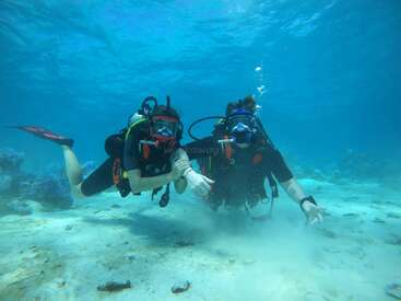 Deux plongeurs sous-marins posent sous l'eau, vêtus de combinaisons noires et d'un équipement complet avec lunettes et bouteilles, explorant un fond marin sablonneux avec des coraux et une eau bleue claire qui les entoure.