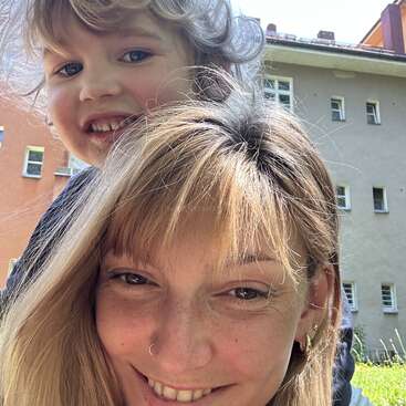 Eine lächelnde Frau mit blonden Haaren und einem Nasenring macht ein Selfie im Freien in Nahaufnahme. Ein Kind mit lockigem Haar umarmt sie von hinten, beide sind glücklich.