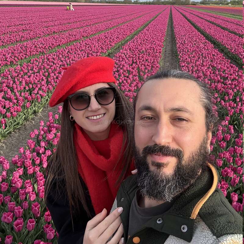 A smiling couple takes a selfie in a vibrant field of blooming pink tulips. The woman wears sunglasses, a red beret, and scarf. Beautiful, colorful scene.