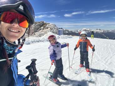 Uma mulher e duas crianças estão sorrindo em uma montanha coberta de neve, vestindo roupas de esqui e capacetes, aproveitando um dia ensolarado de esqui com uma linda vista para as montanhas.