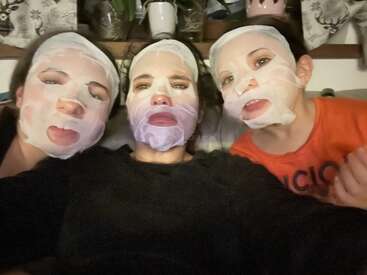Três pessoas estão relaxando juntas, usando máscaras faciais feitas de lençóis brancos. Elas estão sentadas bem pertinho umas das outras, curtindo uma sessão de cuidados com a pele em casa. Detrás delas, dá pra ver vasos de plantas e cortinas estampadas.