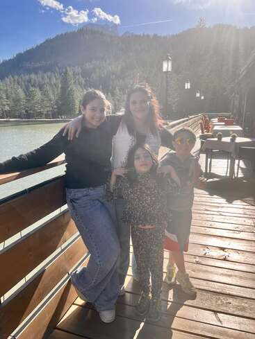 Uma mulher sorridente está em pé com três crianças em um deck de madeira à beira de um lago, cercada por montanhas e pinheiros, aproveitando um dia ensolarado e tranquilo ao ar livre.