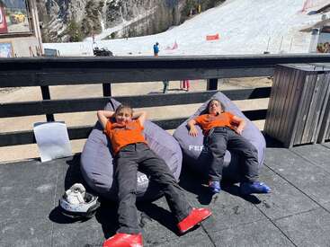 Duas crianças, vestindo camisetas laranja e calças pretas, estão relaxando em pufes ao ar livre, perto de uma encosta coberta de neve, aproveitando o sol, com as botas de esqui e o capacete ao lado.