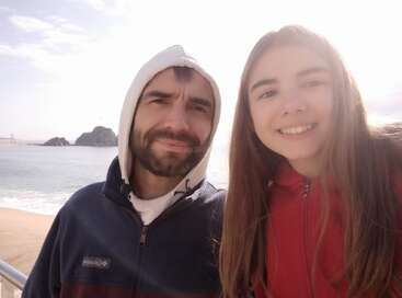 Ein Mann und ein Mädchen in Kapuzenpullis lächeln in der Nähe eines Strandes in die Kamera. Die Sonne scheint hell, im Hintergrund sind die Wellen des Meeres und die Felsformationen zu sehen.