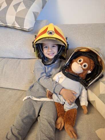 Un enfant souriant est assis sur un canapé, coiffé d'un casque de pompier, et tient dans ses bras un singe en peluche qui porte lui aussi un casque. Tous deux ont l'air heureux et bien au chaud l'un contre l'autre.