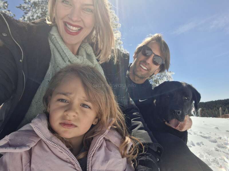 Un selfie de famille joyeux en plein air : une femme souriante, un homme aux lunettes de soleil, une petite fille en veste rose et un chien noir. Une journée ensoleillée, avec des arbres en arrière-plan.