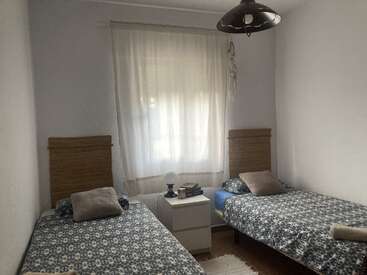 Dieses Bild zeigt ein schlichtes, gemütliches Schlafzimmer mit zwei Einzelbetten, passender gemusterter Bettwäsche, einem gemeinsamen Nachttisch, sanfter Beleuchtung, weißen Vorhängen und dezenten Deko-Akzenten.