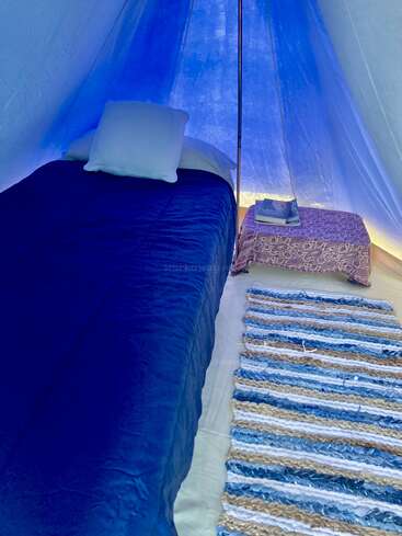 In einem blau beleuchteten Zelt steht ein gemütliches Bett mit weißen Kissen neben einem kleinen Tisch, auf dem Bücher liegen. Ein gestreifter Teppich liegt auf dem Boden und sorgt für Gemütlichkeit.