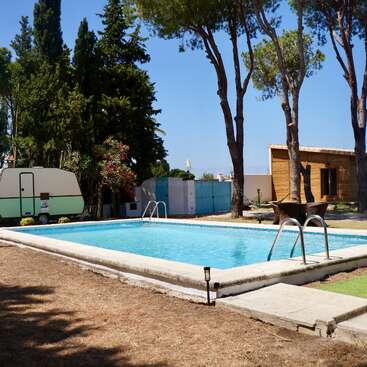 A serene outdoor swimming pool is surrounded by dry grass, tall trees, a wooden cabin, a small green caravan, and clear blue skies, creating a peaceful retreat.