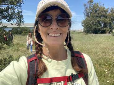 Eine lächelnde Frau mit Zöpfen, die eine Sonnenbrille, eine Mütze und einen Rucksack trägt, macht auf einer sonnigen Wiese ein Selfie, während im Hintergrund ihre Freunde zu sehen sind.