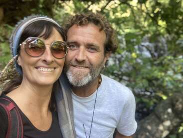 Ein lächelndes Paar posiert draußen für ein Selfie, umgeben von üppig grünen Bäumen. Die Frau trägt eine Sonnenbrille und ein Kopftuch, beide wirken glücklich und entspannt.