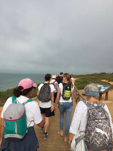 Eine Gruppe von Leuten mit Rucksäcken spaziert auf einem Holzsteg am Meer entlang. Der Himmel ist bewölkt, und grüne Sträucher säumen den Weg und sorgen für eine malerische Aussicht.