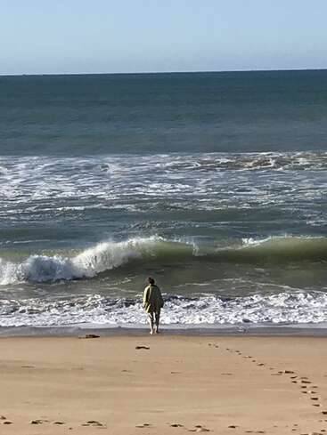 A person in a green garment stands at the edge of the ocean, facing the waves. Fresh footprints trail behind on the golden sandy beach.