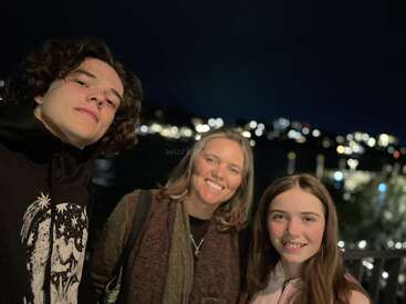 Three people, two young and one adult, smile together outdoors at night. Blurred city lights shine in the dark background, creating a warm, joyful atmosphere.
