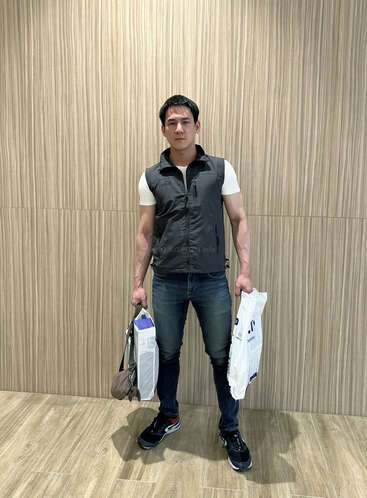 Un homme se tient debout contre un mur à motif bois, vêtu d'un gilet gris, d'un t-shirt blanc, d'un jean et de baskets, tenant des sacs de courses dans les deux mains, l'air sérieux.