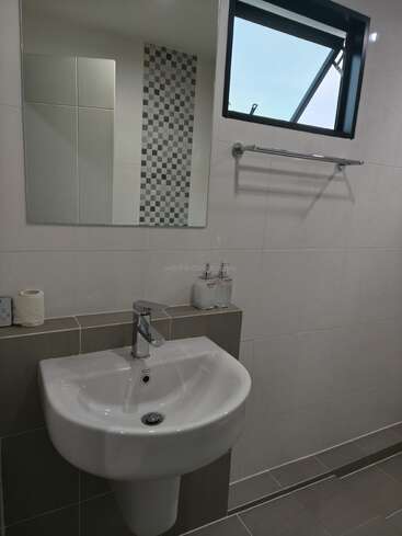Um banheiro moderno tem pia branca, torneira cromada, espelho na parede, pequenos produtos de higiene pessoal, papel higiênico, toalheiro, paredes revestidas com azulejos e uma janela que deixa a luz natural entrar.