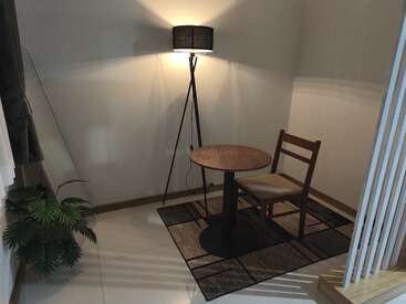 Um canto aconchegante tem uma mesa redonda de madeira, uma cadeira, um abajur e um tapete. Um vaso de planta dá um toque de verde ao lado das janelas com cortinas e da escada.