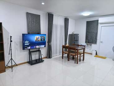 Essa imagem mostra um espaço moderno e arrumado com uma TV, um tripé, mesa de jantar, geladeira, cortinas, iluminação forte e decoração minimalista em piso de azulejo branco.
