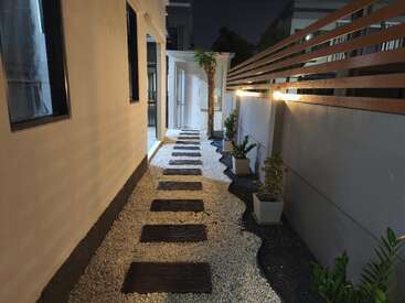 Um caminho estreito ao ar livre à noite, com pedras retangulares, cascalho branco, vasos de plantas, uma cerca de madeira e iluminado por luzes quentes nas paredes.