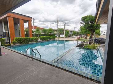 Uma piscina moderna ao ar livre com azulejos azuis está cercada por plantas e prédios urbanos. O céu está nublado e há carros estacionados nas proximidades. Atmosfera tranquila.