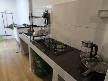 Essa cozinha moderna tem uma bancada preta, fogão a gás, chaleira, escorredor de pratos, utensílios, pia, botijão de gás verde e produtos de limpeza contra paredes com azulejos brancos.