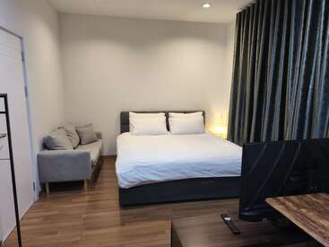 Essa imagem mostra um quarto moderno e arrumado com uma cama grande, sofá cinza, piso de madeira, TV, decoração minimalista, paredes brancas e cortinas escuras. Ambiente aconchegante.