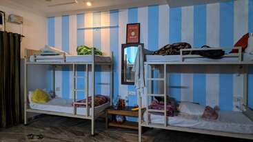 A imagem mostra um quarto com duas beliches de metal, cada uma com roupa de cama e mochilas. As paredes são listradas de azul e branco, há um espelho e uma mesinha.