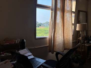 Um aconchegante espaço de trabalho com um laptop, um caderno e uma cadeira fica junto a uma janela ensolarada com cortinas transparentes, com vista para campos verdejantes e árvores lá fora.