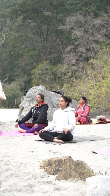 Três mulheres estão sentadas de pernas cruzadas em tapetes de ioga ao ar livre, meditando tranquilamente. Elas estão rodeadas por árvores, pedras e areia, criando um ambiente calmo e natural.