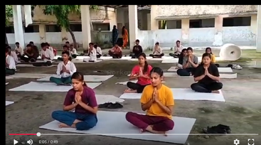 Um grupo de alunos está sentado de pernas cruzadas em esteiras ao ar livre, com os olhos fechados e as mãos em posição de oração, participando de uma sessão coletiva de meditação ou ioga na escola.