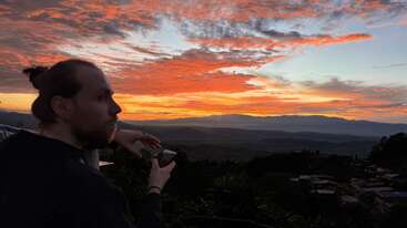 A person with a bun gazes at a vibrant, colorful sunset over distant mountains, holding a phone, surrounded by scenic natural beauty and silhouetted rooftops.