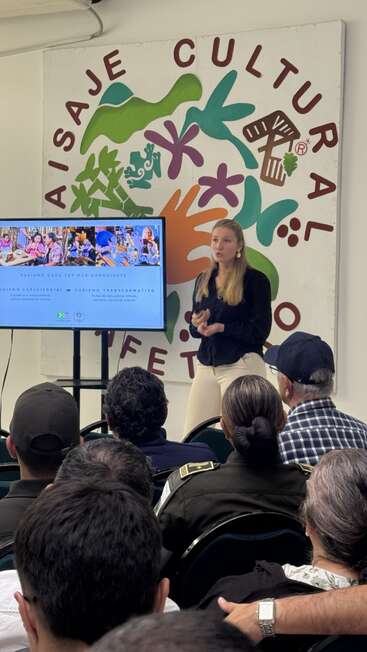 A woman gives a presentation about conscious, transformative tourism to an attentive audience, with colorful cultural artwork and a digital screen displaying information and photos behind her.