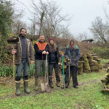 The image depicts four individuals standing in a wooded area, each holding gardening tools, with a pile of logs and trees visible in the background.