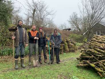 The image depicts four individuals standing in a wooded area, each holding gardening tools, with a pile of logs and trees visible in the background.