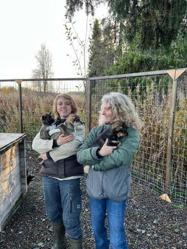 Deux femmes se tiennent à l'extérieur, chacune tenant un chiot, avec un grillage et des arbres en arrière-plan, dans un cadre extérieur serein.