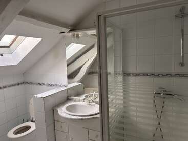 Il s'agit d'une petite salle de bains carrelée de blanc, dotée d'un plafond en pente, d'une lucarne, de toilettes, d'un lavabo avec meuble, d'un grand miroir et d'une cabine de douche en verre.