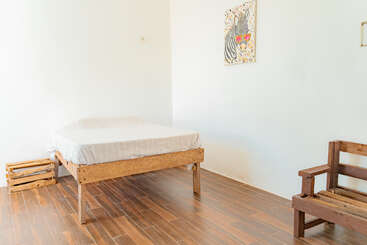 A minimalist bedroom with a simple wooden bed, white bedding, a wooden crate, wooden bench, wooden floor, plain white walls, and a zebra-themed artwork.