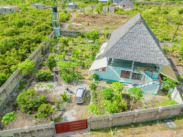 This image shows an aerial view of a house with a thatched roof, surrounded by a lush garden, vegetation, boundary wall, water tower, and a parked car.