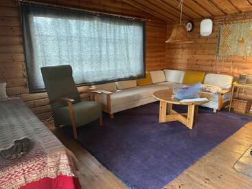 Esta acogedora habitación de madera cuenta con un sillón verde, un sofá modular blanco, una mesa redonda de madera, una alfombra morada, una ventana con cortinas, una pantalla de lámpara tejida y un tapiz decorativo en la pared.