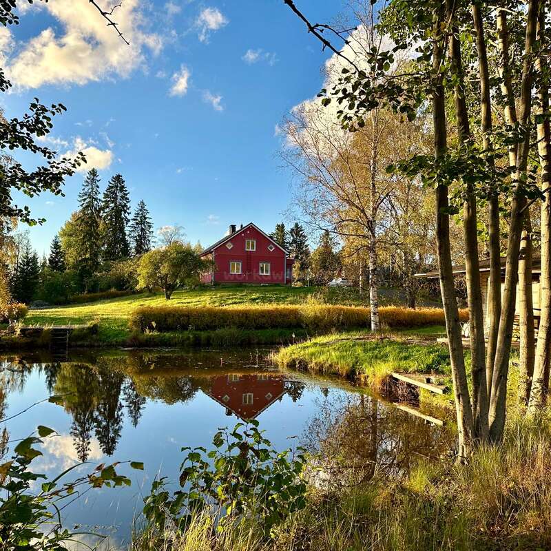 Una encantadora casa roja se alza sobre una colina cubierta de hierba, rodeada de árboles, y su reflejo se refleja con belleza en un tranquilo estanque bajo un cielo azul y despejado.