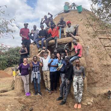 Un grupo heterogéneo de personas posa feliz frente a un edificio de tierra, celebrando el trabajo en equipo y los logros, rodeados de naturaleza y cielos despejados, exudando unidad y alegría.