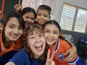 Una alegre foto de grupo en la que aparecen una profesora sonriente y cuatro jóvenes alumnos con uniformes naranjas y azules, posando juntos en un aula muy iluminada y haciendo el signo de la paz.